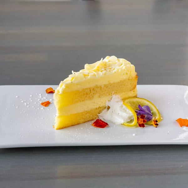 A slice of lemon cake with white chocolate shavings, whipped cream, lemon slices, and edible flowers.
