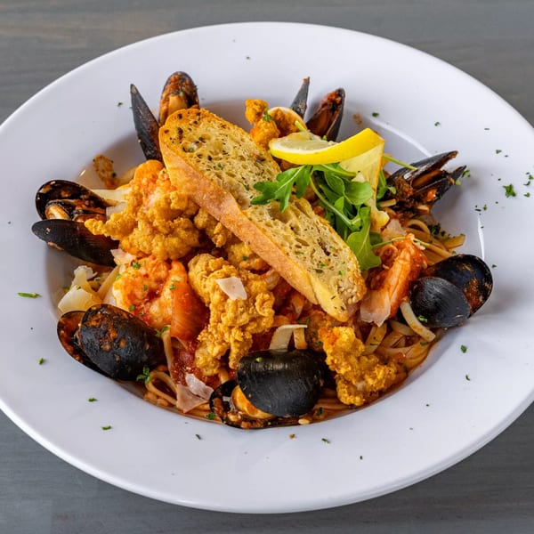 Seafood pasta with mussels, fried calamari, shrimp, garlic bread, and lemon on a white plate.