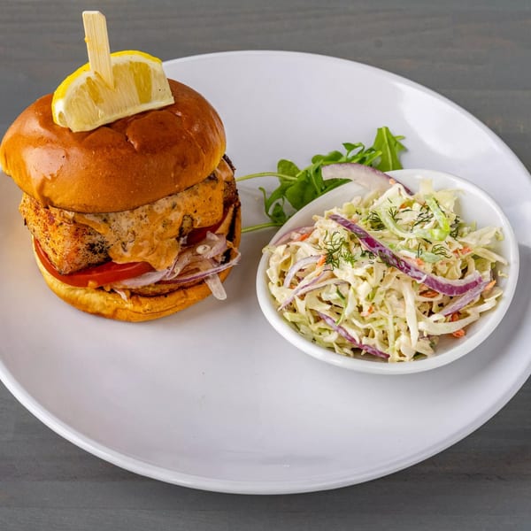 Grilled fish sandwich with a lemon wedge, tomato, onion, and a side of coleslaw.